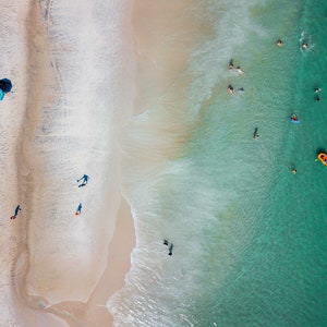 May include: An aerial view of a sandy beach with turquoise water. People are swimming and floating in the water. A blue beach umbrella is on the sand.