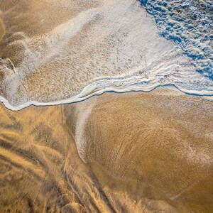 May include: An aerial view of a sandy beach with a wave breaking on the shore. The wave is white with blue water and the sand is a light brown color.