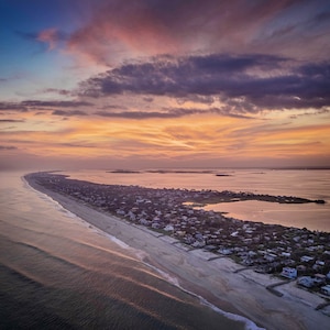 May include: Aerial view of a coastal town at sunset. The sky is a blend of blue, orange, and purple hues, with clouds. A long stretch of beach borders the town, with houses and buildings. The ocean is calm, reflecting the colors of the sky.