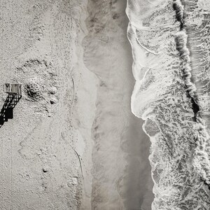 May include: Aerial black and white photograph of a beach scene. A lifeguard stand casts a long shadow on the sand. Waves crash along the shoreline, creating a textured pattern. The image captures the natural beauty of the coast.
