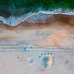 May include: Aerial view of a beach with green ocean waves washing onto a sandy shore. A blue umbrella and beach chairs are on the sand. Large footprint impressions are visible in the sand, suggesting a beach theme.