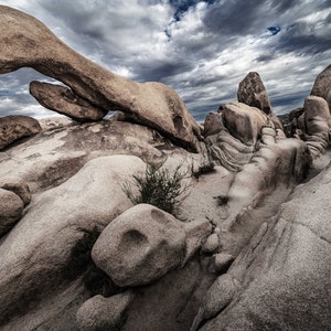 May include: A large rock formation with a natural archway in the desert. The rock is a light gray color and the sky is a cloudy blue.