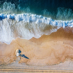 May include: An aerial view of a sandy beach with a lifeguard stand and waves crashing on the shore. The water is a deep blue and the sand is a light brown.