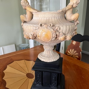 May include: A decorative urn with cherub figures, floral accents, and a central medallion. The urn is cream-colored and sits atop a black pedestal. The overall design is ornate and classical, suitable for home decor.