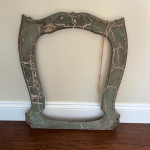 May include: An ornate, weathered wooden mirror frame featuring a distressed sage green finish with visible cracks and exposed wood grain. The frame has a curved, decorative top crest and a wide, open center, with a thin piece of twine hanging down.