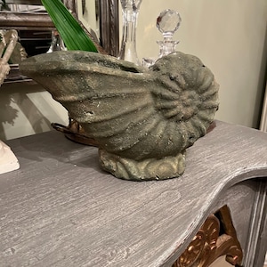 May include: A weathered, green-toned concrete seashell sculpture. The nautilus-shaped piece has a textured surface and sits on a gray table. Decorative glass bottles are in the background. The sculpture is a unique home decor item.
