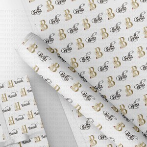 May include: White wrapping paper with a repeating pattern of the letter "B" in gold with the name "Bella" in a black script font. A wrapped gift with the name "Hannah" is also visible.
