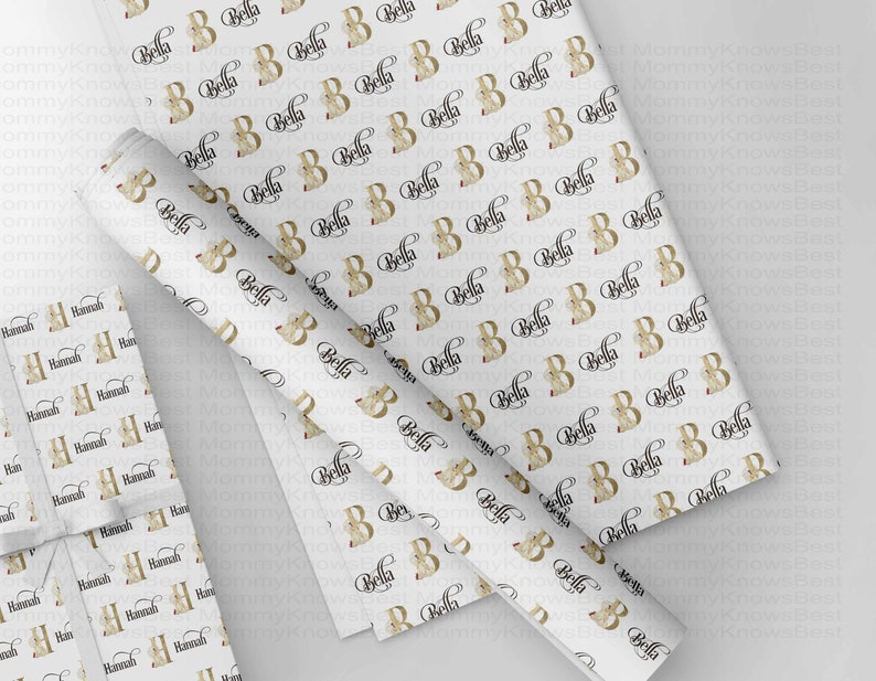 May include: White wrapping paper with a repeating pattern of the letter "B" in gold with the name "Bella" in a black script font. A wrapped gift with the name "Hannah" is also visible.