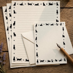 Op de afbeelding: Een set cr&egrave;mekleurig briefpapier met een rand van zwarte katten en pootafdrukken. De set bevat meerdere gelinieerde vellen, een kleinere ongelinieerde kaart, een bijpassende envelop en een houten kroontjespen om mee te schrijven.