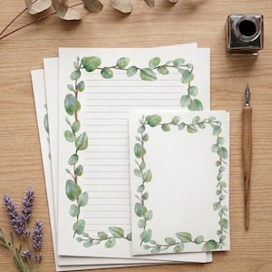 Puede incluir: Juego de papelería con papel rayado y en blanco, cada uno con un borde de hojas de eucalipto en acuarela. Un pequeño bloc de notas, un tintero y una pluma están dispuestos sobre una superficie de madera, junto con ramitas de eucalipto y lavanda secas. El papel tiene un diseño botánico delicado.