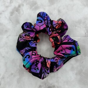 Könnte beinhalten: Ein lila, rosa und blauer Haargummi mit einem Würfelmuster. Die Würfel sind schillernd und haben schwarze Zahlen.