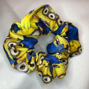 May include: A yellow and blue scrunchie featuring a Minion character print. The scrunchie is made of fabric and has a gathered design.