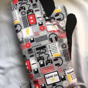 May include: A gray oven mitt with a black heat-resistant lining. The mitt features a repeating pattern of video game and computer icons, including joysticks, headphones, and floppy disks. The text "GAME ON" appears multiple times on the mitt.