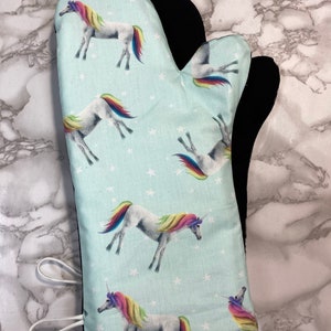 Puede incluir: Un guante para horno azul claro con un forro negro resistente al calor. El guante presenta un patrón repetitivo de estrellas blancas y unicornios coloridos con crines y colas de arcoíris.