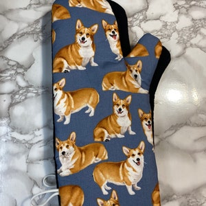 May include: A blue oven mitt with a pattern of brown and white corgis. The mitt has a black heat-resistant lining and a white loop for hanging.
