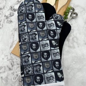 May include: A black oven glove with a square pattern featuring black and white horror movie character portraits. The glove has a black interior and a white trim. Wooden spoons and measuring spoons are in the background.
