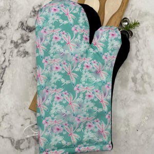 May include: A green oven mitt with a dragonfly and floral pattern. The mitt is lined with black fabric.