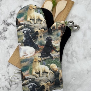 Könnte beinhalten: Ein weißer Ofenhandschuh mit einer schwarzen hitzebeständigen Innenauskleidung zeigt einen farbenfrohen Druck von Labrador Retrievern in einer Waldlandschaft.