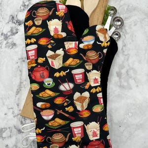 May include: A black oven mitt featuring a colorful pattern of various Asian-inspired food items, including takeout boxes, teapots, spring rolls, fortune cookies, and bowls of noodles. The mitt has a solid black interior lining and a white hanging loop.