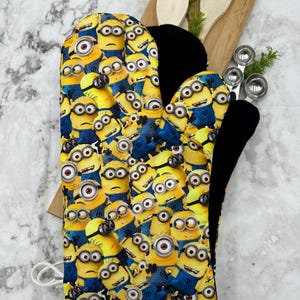 Puede incluir: Un guante de horno amarillo con un forro negro resistente al calor. El guante está cubierto con un patrón de Minions de dibujos animados.