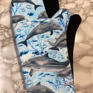 Peut inclure: Une manique de four bleue et grise avec un motif de dauphins et de poissons. La manique est doublée d'une couche noire résistante à la chaleur.