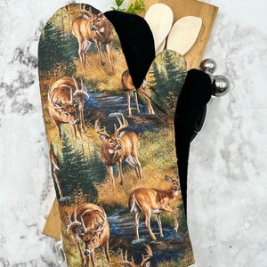 Peut inclure: Une manique de four marron et noire avec une bordure blanche. La manique présente un motif répété de cerfs dans un décor forestier.