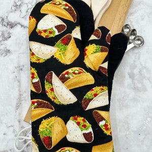 May include: Black oven mitt with a white and yellow pattern of tacos. The tacos have fillings of lettuce, cheese, and meat.