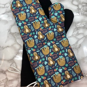 May include: A pair of oven mitts with a black heat-resistant lining and a blue cotton outer layer. The cotton layer features a repeating pattern of brown sloths hanging from branches with the text "Born to be Mild" printed on each sloth.