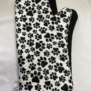 Puede incluir: Un guante de cocina blanco con un estampado de huellas de patas negras. El guante tiene un ribete negro y un lazo para colgar. Este accesorio de cocina está diseñado para proteger las manos del calor.