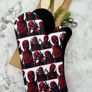 May include: A white oven mitt with a repeating pattern of Deadpool's face. The oven mitt is folded in half and has a white trim around the edges.