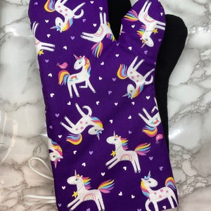 Puede incluir: Manopla de horno morada con un estampado de unicornios blancos con crines y colas de arcoíris, y pequeños corazones morados. La parte trasera de la manopla es negra. Se adjunta un lazo blanco para colgar.