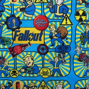 May include: A blue fabric featuring a repeating pattern of characters and symbols from the Fallout video game series. The design includes the word "Fallout", Vault Boy, Nuka Cola logos, and hazard symbols.