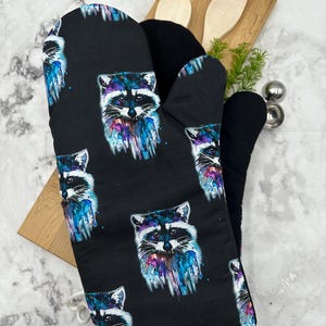 May include: Black oven mitt with a watercolor raccoon print. The raccoon is depicted in shades of blue, purple, and white against a black background.