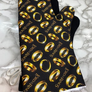 May include: Black oven mitt with a white trim and a repeating pattern of gold rings and the words "The Lord of the Rings" on a black background.