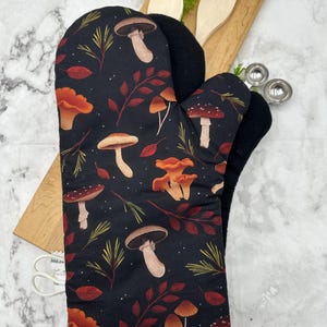 May include: An oven mitt featuring a black fabric base with a repeating pattern of various mushrooms, red and brown leaves, and green pine needles. The mitt has a white trim at the opening and a black interior lining, designed for kitchen use.