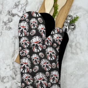 May include: Black oven mitt with a repeating pattern of a white hockey mask with red blood splatters. The mask is worn by a figure holding a machete. The oven mitt has a black lining and a white string.