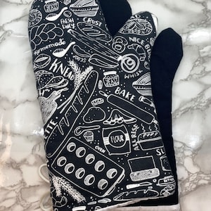 May include: A black oven mitt featuring a white line-art pattern of baking-related illustrations and text including the words KNEAD, BAKE, FLOUR, YEAST, CRUST, and NICE BUNS. The mitt has a solid black back and a white hanging loop at the base.