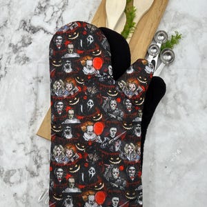 May include: A black oven mitt with a white trim features a colorful pattern of horror movie characters and jack-o-lanterns.