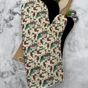 May include: A cream-colored oven mitt with a black heat-resistant lining. The mitt features a repeating pattern of cats with wings, flowers, and the word "MEOW" in a vintage tattoo style.