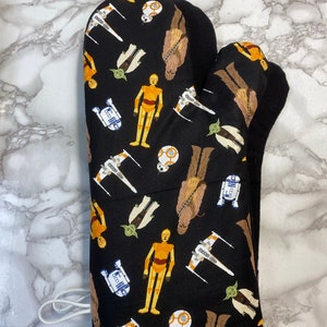 Puede incluir: Manopla de horno negra con un estampado de Star Wars con personajes como Chewbacca, R2-D2, C-3PO, Yoda y cazas X-Wing.