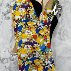 May include: An oven glove with a vibrant print of The Simpsons characters. The design features Homer, Bart, and other characters in a repeating pattern. The glove has a black interior and a white hanging loop. The Simpsons logo is visible.