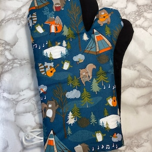May include: A blue oven mitt with a camping theme. The mitt features a pattern of animals playing music in a forest setting, including a bear, raccoon, owl, and squirrel. The mitt has a black heat-resistant lining.