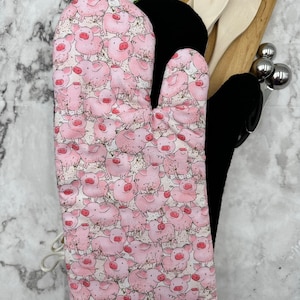 Könnte beinhalten: Ein rosa Ofenhandschuh mit einem Muster aus rosa Schweinen auf weißem Hintergrund. Der Handschuh hat eine schwarze hitzebeständige Schicht auf der Rückseite.
