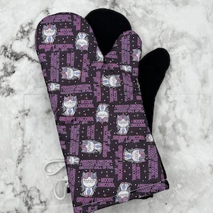 Puede incluir: Un par de guantes de horno negros con un patrón de unicornios morado y blanco. El patrón incluye el texto "Annoyed Unicorn, Touchy Unicorn, Grumpy Ball of Fur, Moody Unicorn, Grumpy Unicorn".
