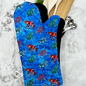 May include: Blue oven mitt with a pattern of colourful frogs on a blue background. The mitt is made of fabric and has a black lining.
