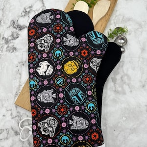 May include: A black oven mitt featuring a colourful pattern of Star Wars characters and symbols in a sugar skull style. The design includes Darth Vader, Boba Fett, and droids in shades of orange, blue, yellow, and white, with a white trim at the base.