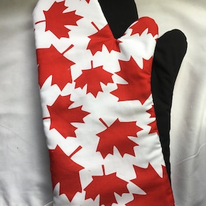 Puede incluir: Manopla de horno roja y blanca con un patrón de hojas de arce. La manopla es negra en la parte posterior.