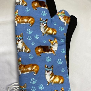 Puede incluir: Una manopla de horno azul con un patrón de corgis con gafas de sol. Los corgis son marrones, negros y blancos. La manopla tiene un forro negro resistente al calor y un lazo blanco para colgar.