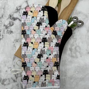 May include: A white oven mitt with a colorful cat print. The mitt is lined with black fabric and has a white loop for hanging.