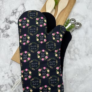 Può includere: Un guanto da forno nero con un motivo ripetuto di ghirlande floreali rosa, gialle e verdi. All'interno di ogni ghirlanda, il testo recita "Fuck this Shit" o "Shut the fuck up" in un carattere corsivo nero. Il guanto ha un bordo bianco.